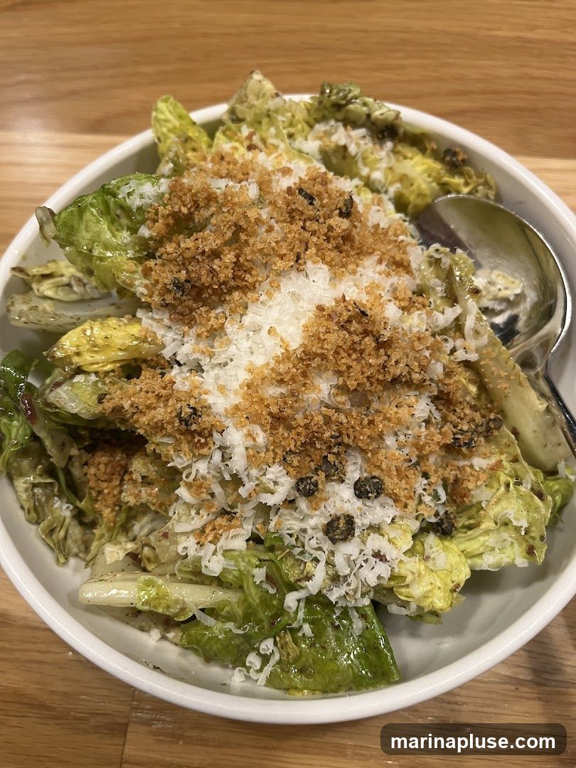 A close-up shot of a roasted romaine salad in a white bowl. The salad is topped with shredded parmesan cheese, breadcrumbs, and capers. A spoon is visible on the right side of the bowl.