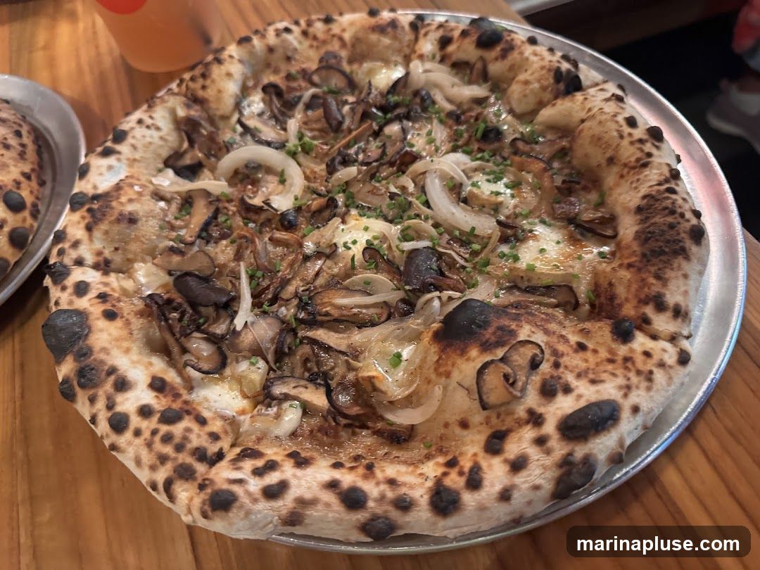 A close-up shot of a mushroom pizza with a charred crust, served on a metal tray. The pizza is topped with various mushrooms, onions, and herbs. Another pizza is partially visible on the left. A drink in a clear cup with an orange tint is in the upper left corner.