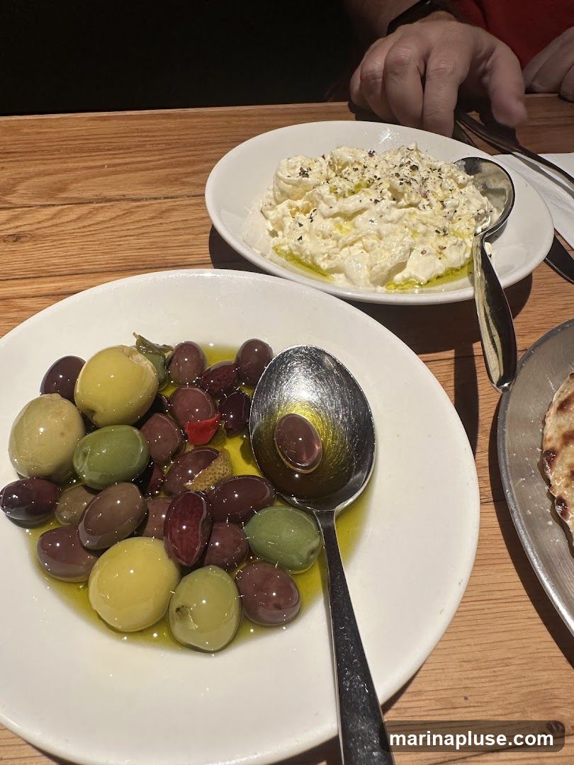 A close-up shot of two white bowls on a wooden table. The bowl in the foreground contains a variety of olives in different colors and sizes, swimming in olive oil. A spoon rests in the bowl. The bowl in the background holds a creamy white dip, sprinkled with herbs and drizzled with olive oil. A person's hand is visible in the upper right corner of the image.
