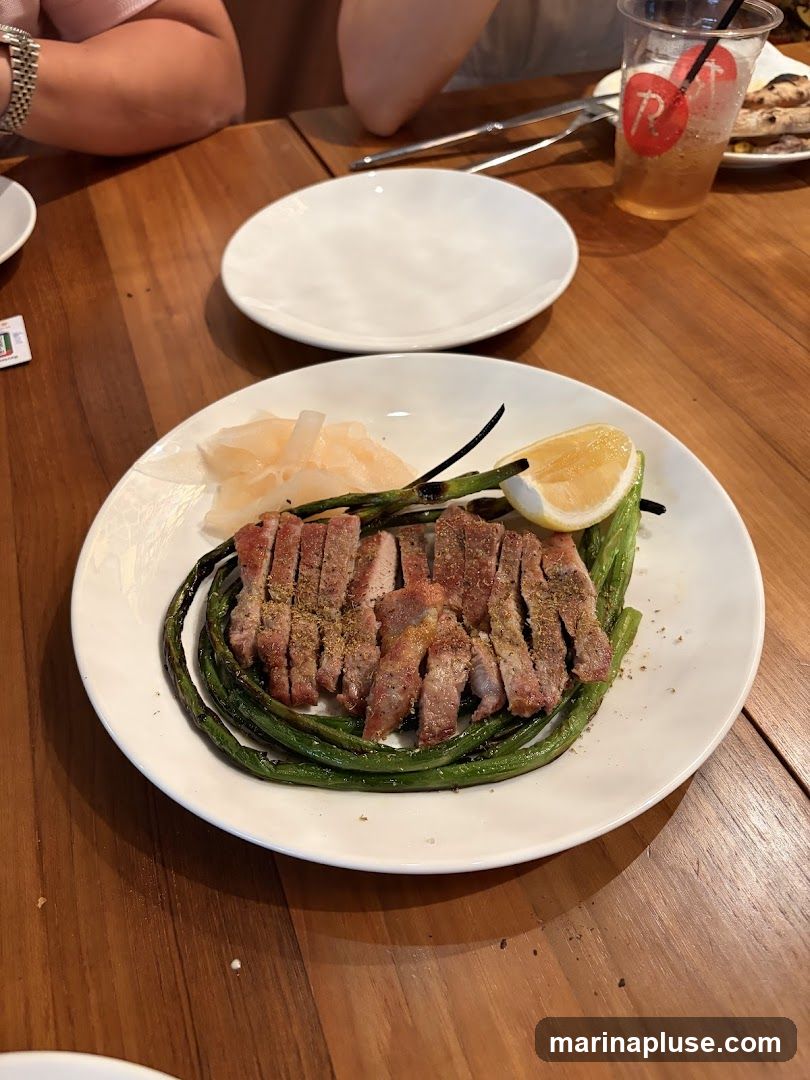 A plate of sliced pork is served with grilled green beans and a lemon wedge. Pickled ginger is also on the plate. The meal is on a wooden table, with a clear plastic cup with a red logo and a straw visible in the background.