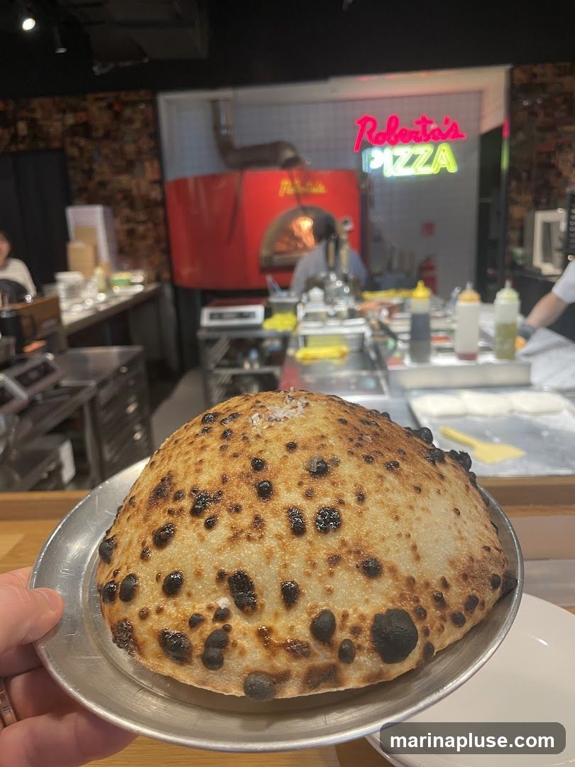 A close-up of a perfectly baked pizza dough ball, lightly sprinkled with salt, sits on a metal plate. In the background, a busy pizza kitchen is visible with a red pizza oven and a neon sign that reads "Roberta's PIZZA".