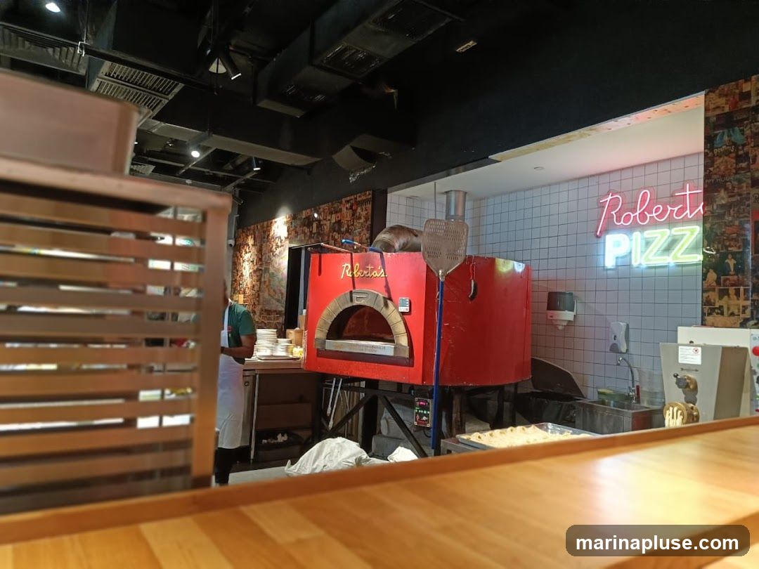 A view of a pizza restaurant's interior, featuring a bright red pizza oven with the name "Roberta's" visible. Above the oven, a neon sign spells out "PIZZ". The walls are tiled, and a person in an apron stands near a stack of plates. A wooden counter is in the foreground.