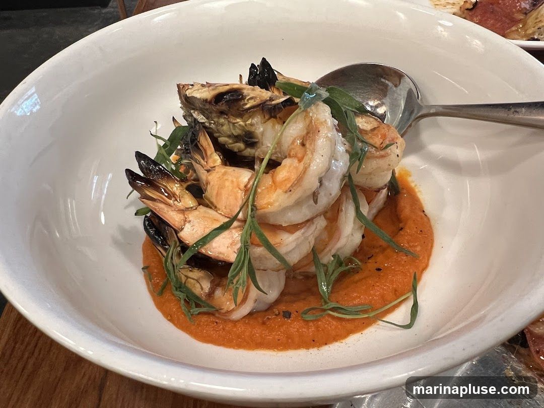 A white bowl filled with grilled shrimp served on a bed of orange sauce and garnished with fresh herbs. A spoon rests in the bowl.