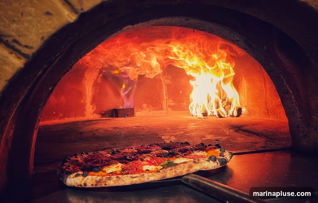 A close-up shot shows a pizza being cooked in a traditional wood-fired oven. Flames are visible in the background, illuminating the interior of the oven. The pizza, with its crust slightly charred and toppings visible, is positioned on a metal peel in the foreground.