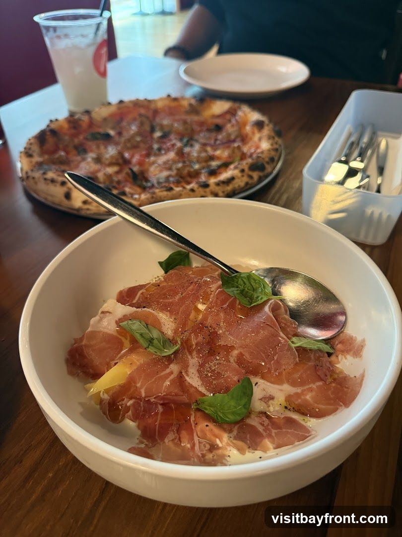A close-up view of a white bowl filled with thinly sliced prosciutto, garnished with fresh basil leaves and a drizzle of olive oil. Behind it, a pizza with a crispy crust and various toppings is visible, along with a clear plastic cup containing a beverage and a white container holding cutlery. The setting appears to be a restaurant table.