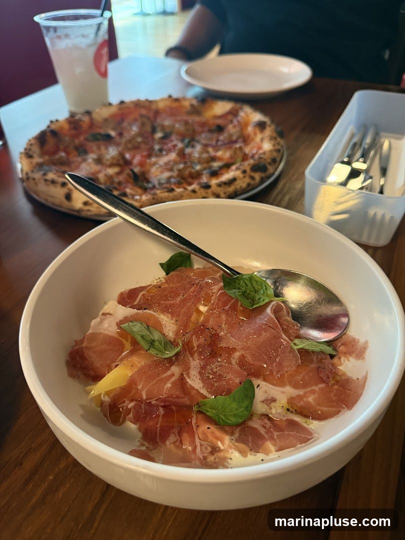 A close-up view of a white bowl filled with thinly sliced prosciutto, garnished with fresh basil leaves and a drizzle of olive oil. Behind it, a pizza with a crispy crust and various toppings is visible, along with a clear plastic cup containing a beverage and a white container holding cutlery. The setting appears to be a restaurant table.