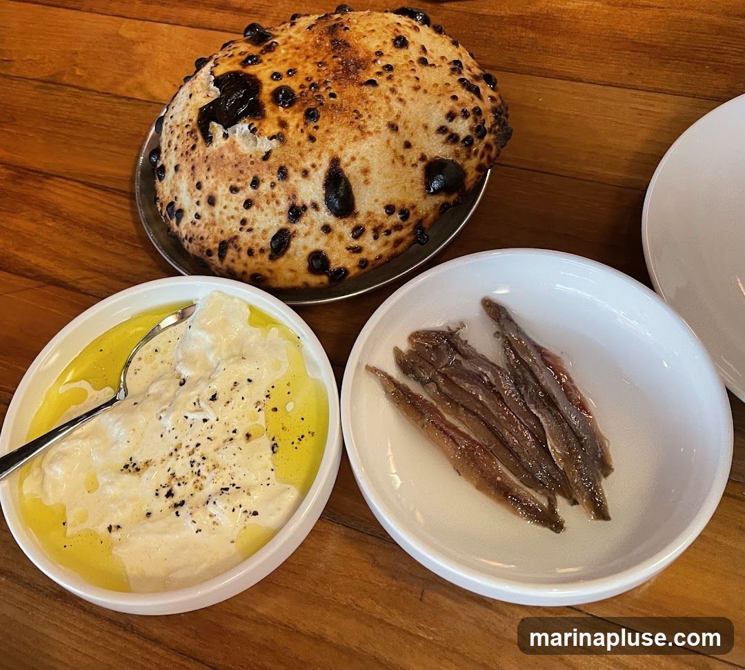 A round, charred flatbread sits on a metal plate. To its left, a white bowl contains a creamy dip with a spoon, drizzled with olive oil and sprinkled with black pepper. To the right, a white bowl holds a serving of anchovies. The food is arranged on a wooden table.