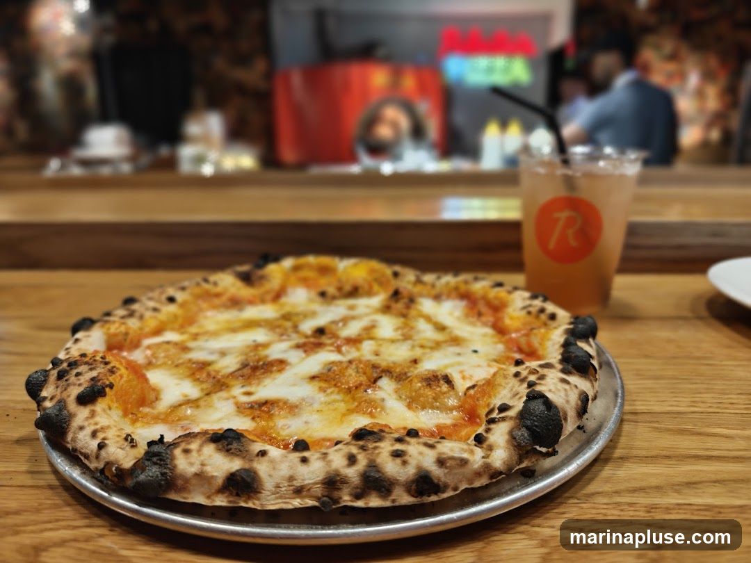 A close-up shot of a pizza and a drink on a wooden table. The pizza has a charred crust and is topped with cheese and tomato sauce. The drink is in a clear plastic cup with a straw and has a logo with the letter 'R' inside a circle. In the background, there is a blurred view of a restaurant interior with neon lights and people.