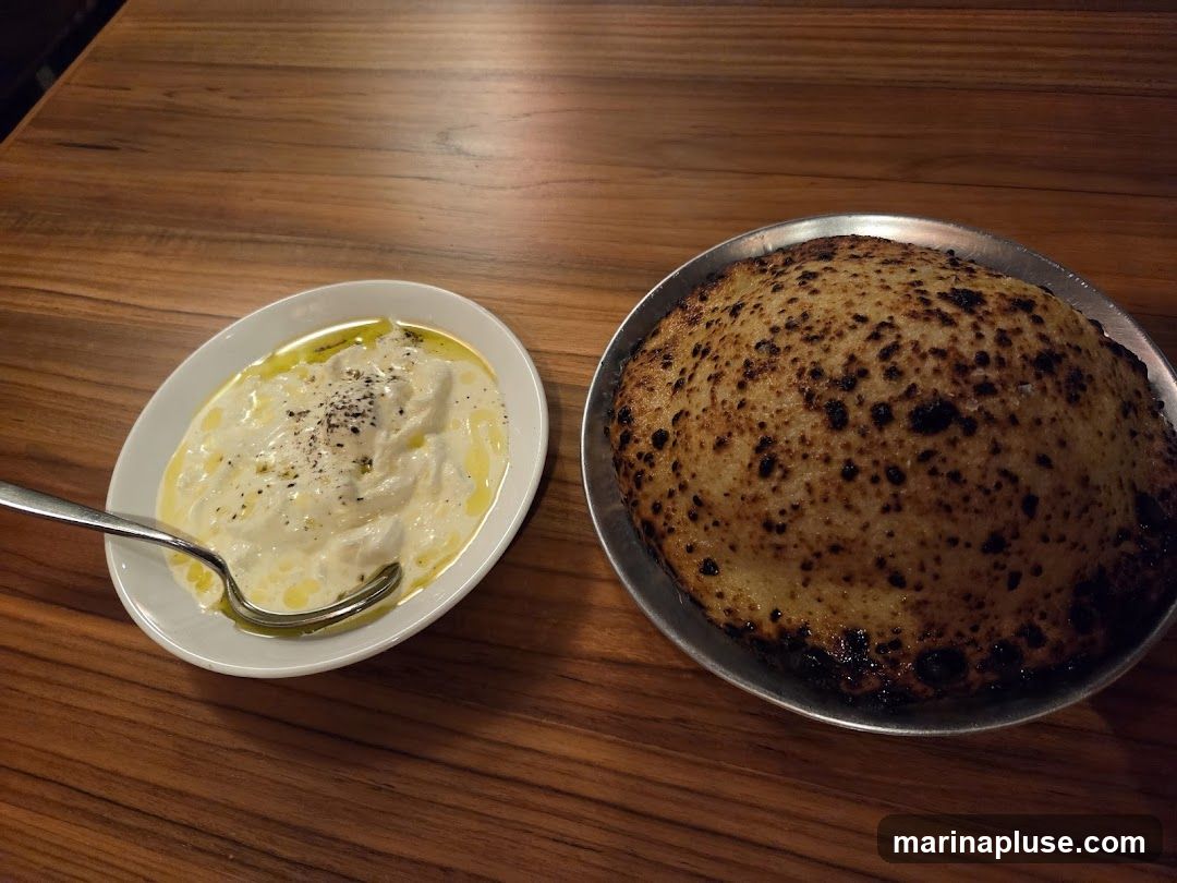 A white oval dish filled with a creamy white dip, drizzled with olive oil and sprinkled with black pepper, sits next to a round, flatbread with charred spots, served on a metal plate. Both are placed on a wooden table.