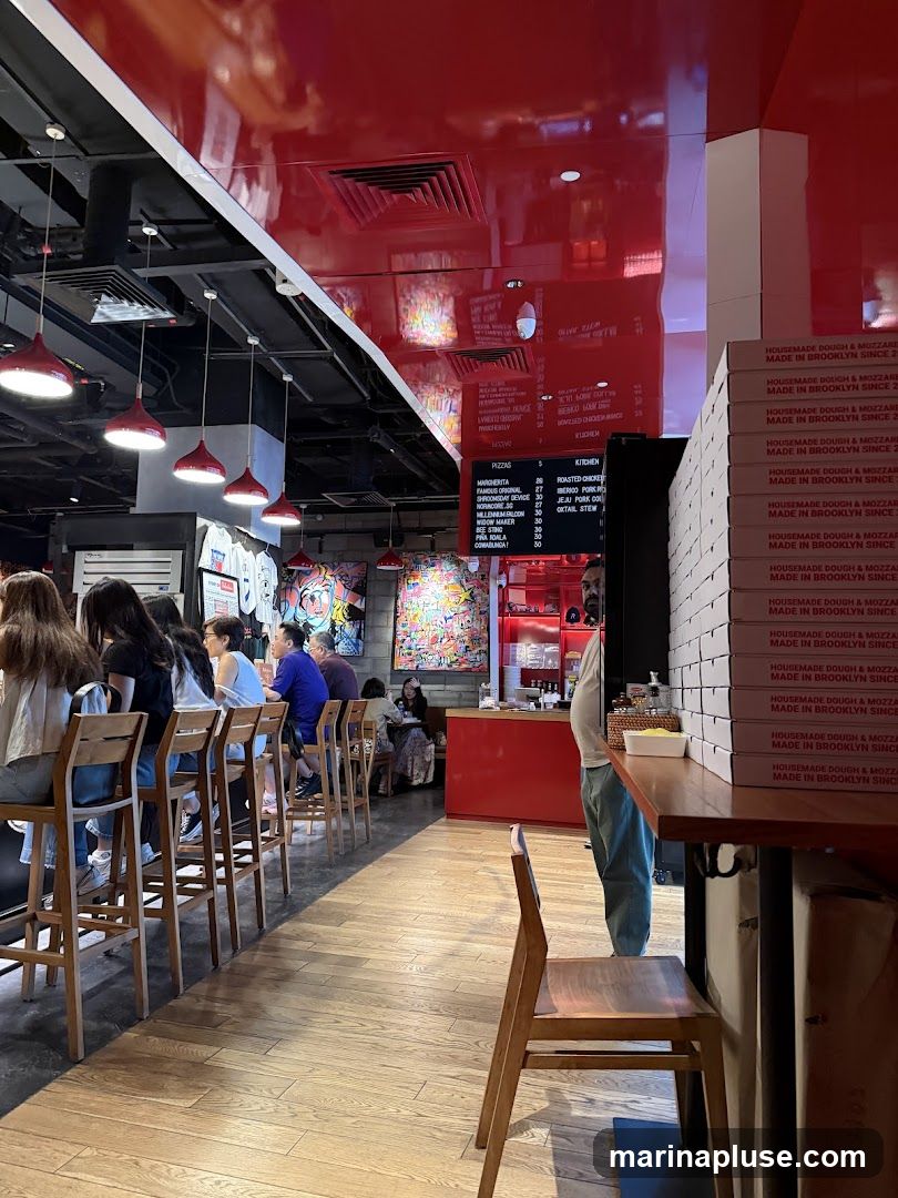 An interior view of a pizza restaurant with a long counter seating area. Several patrons are seated at the counter. A menu board is visible on the wall, displaying pizza and kitchen items. Stacks of pizza boxes are visible on a table in the foreground. The decor includes red accents and colorful artwork.