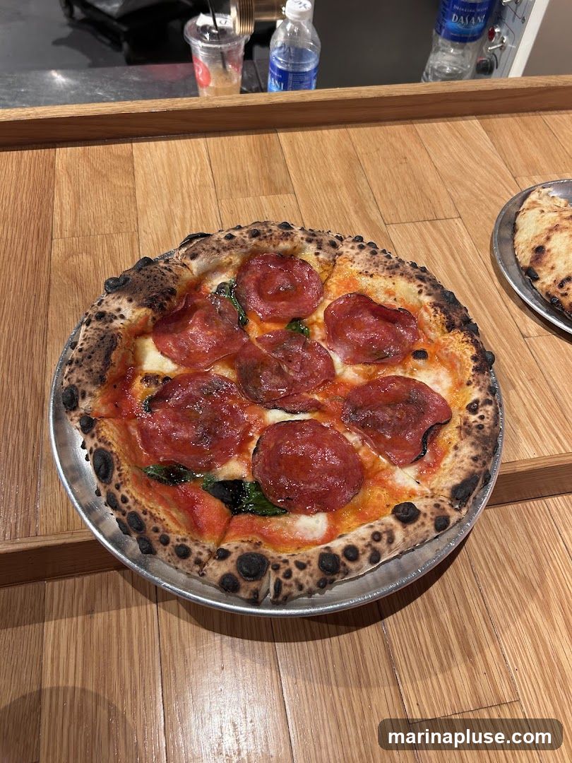 A close-up shot of a pepperoni pizza with a charred crust, served on a metal plate. The pizza has a tomato sauce base, melted cheese, and slices of pepperoni. There are also some green leaves, likely basil, scattered on top. In the background, there are bottles of water and a cup with a straw on a wooden counter. Another flatbread or pizza is visible on a plate to the right.