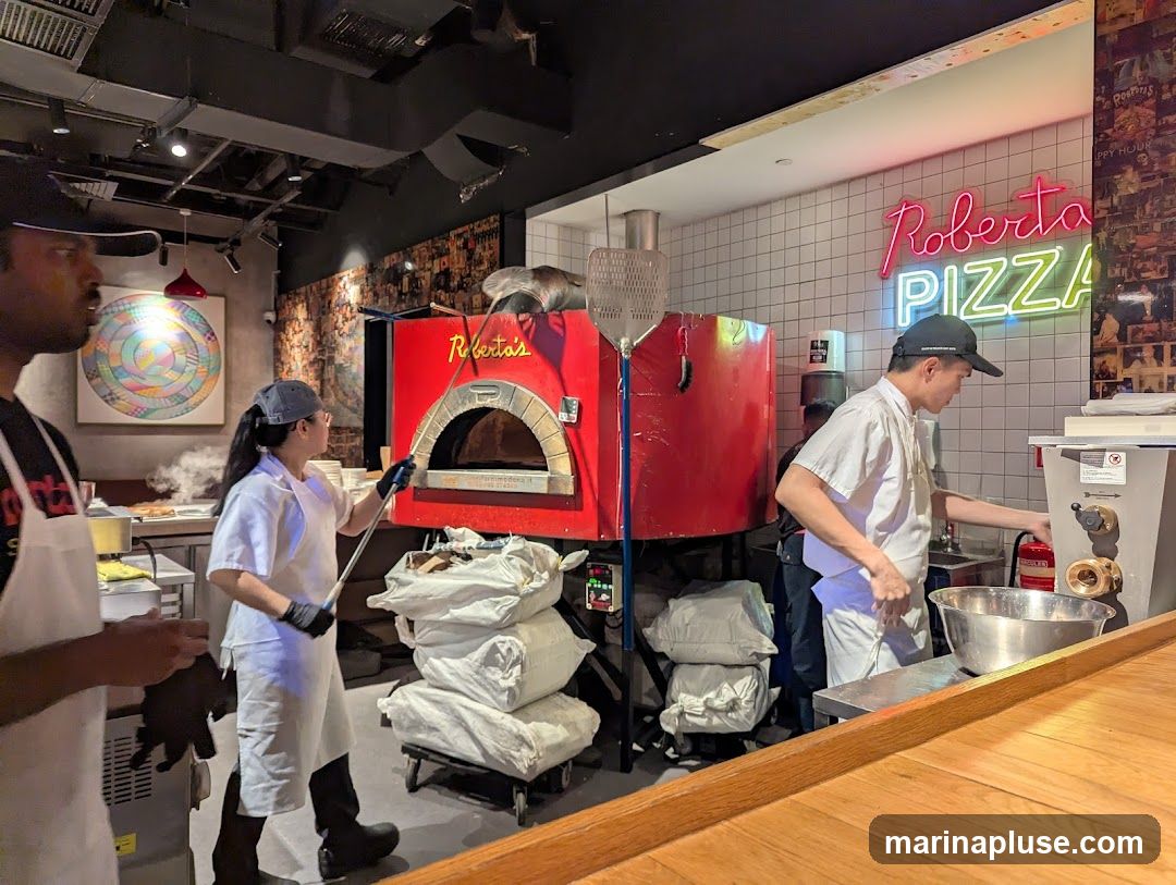 A busy pizza restaurant kitchen with staff preparing food. A red pizza oven is visible, along with a neon sign that reads "Roberto's PIZZA". One staff member is using a pizza peel to tend to the oven, while another is working at a counter with a large metal bowl. Bags of flour are stacked nearby.