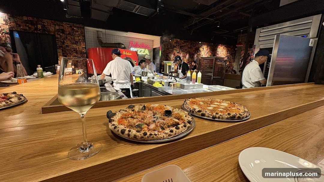 Two pizzas and a glass of white wine are on a wooden counter in front of a busy pizza kitchen. Chefs are visible working behind the counter, preparing food. A neon sign that says "Roberto's PIZZA" is visible in the background. The walls are decorated with a collage of images.